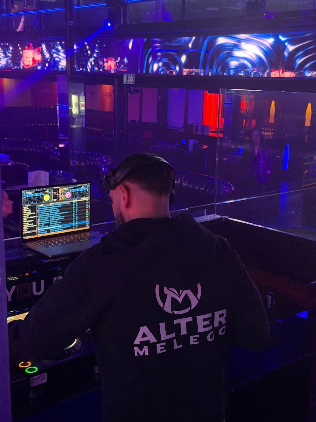 Alter Melego from behind at the decks, logo on his hoodie, club lights ahead